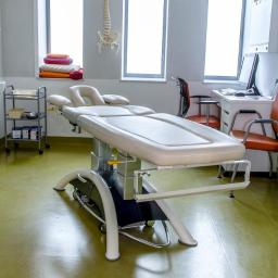 Arsmedis - Jasny gabinet z regulowanym stołem do masażu, szkieletem kręgosłupa na ścianie i logo ArsMedis nad oknem.