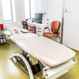 Arsmedis - Jasne pomieszczenie z białym stołem do masażu, w tle biurko z komputerem, model kręgosłupa na ścianie i wózek z akcesoriami.