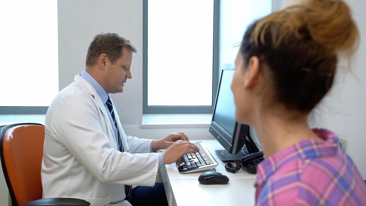 Lekarz w białym kitlu wpisuje dane pacjentki do komputera, obok stoi monitor i telefon stacjonarny, w tle okno.