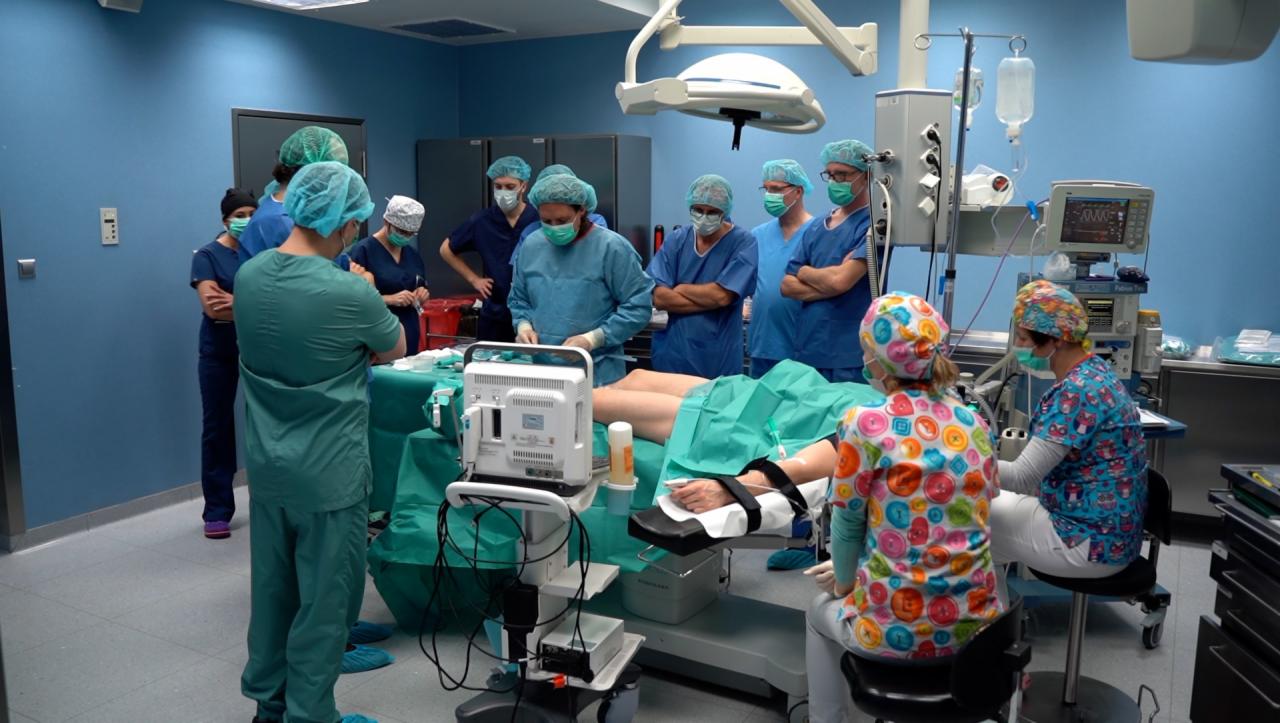 Sala operacyjna z zespołem medycznym w trakcie zabiegu, widoczny pacjent na stole operacyjnym, aparatura medyczna i lampa operacyjna, personel w strojach ochronnych i kolorowych czapkach.