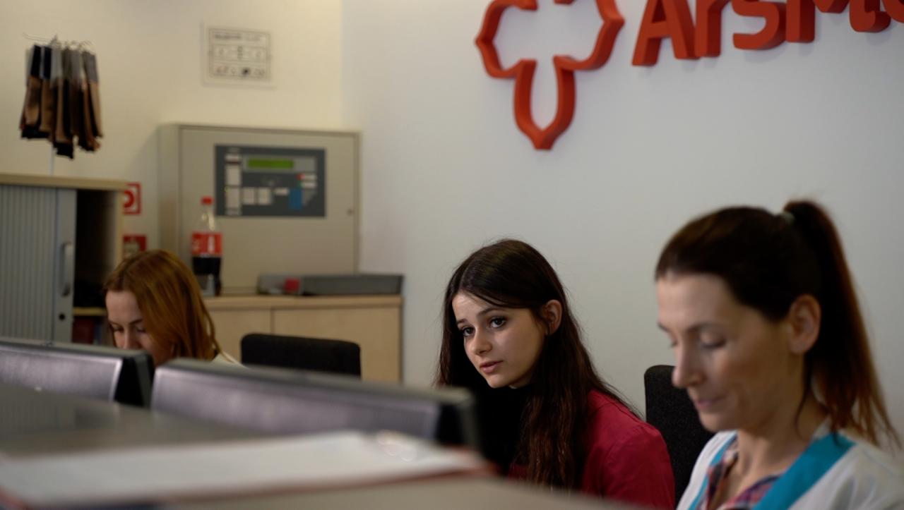 Recepcja placówki medycznej: trzy kobiety siedzą za biurkiem, w tle logo firmy i szafka z dokumentami.