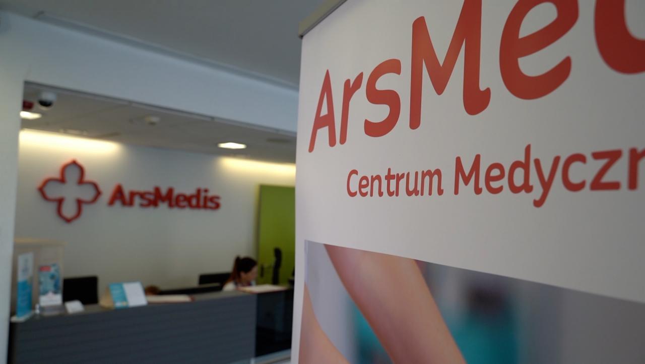 Recepcja Centrum Medycznego ArsMedis z widocznym logo na ścianie i banerem reklamowym z częścią ciała w tle.