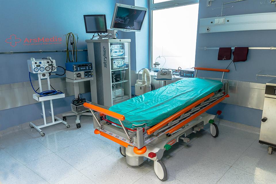 Sala szpitalna z łóżkiem transportowym, sprzętem medycznym Stryker i monitorami, oświetlona światłem dziennym z okna.