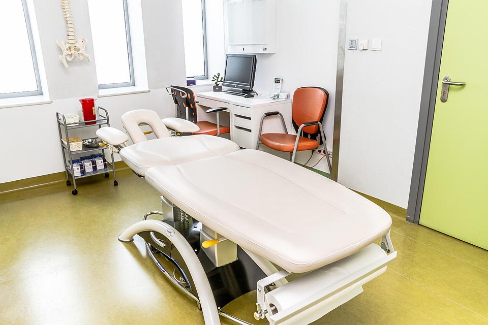 Jasne pomieszczenie z białym stołem do masażu, w tle biurko z komputerem, model kręgosłupa na ścianie i wózek z akcesoriami.