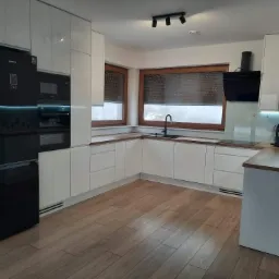 Nowoczesna kuchnia z białymi, lakierowanymi frontami, drewnianymi blatami, czarnym sprzętem AGD i podłogą z paneli imitujących drewno.