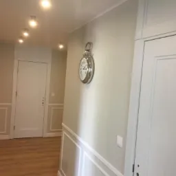 Korytarz po remoncie z panelami ściennymi w stylu angielskim, ozdobiony dużym, srebrnym zegarem kieszonkowym na ścianie i punktowym oświetleniem sufitowym.
