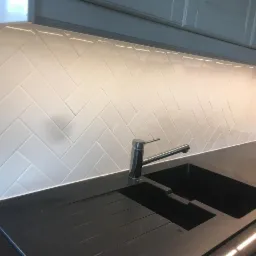 Białe, błyszczące płytki ułożone w jodełkę na ścianie nad czarnym, dwukomorowym zlewem i chromowaną baterią w kuchni. Pod szafkami górnymi widoczne oświetlenie LED.