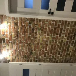 Ceglana ściana z widocznymi fugami, oświetlona dwoma punktowymi lampami, sąsiadująca z białymi, drewnianymi elementami zabudowy z niebieskimi akcentami.