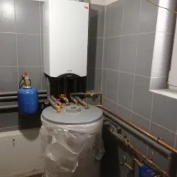 Nowoczesny, biały piec grzewczy wiszący na szarej, kafelkowanej ścianie, podłączony do instalacji i zbiornika ciepłej wody owiniętego folią ochronną.