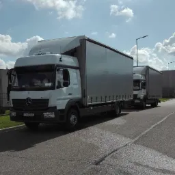 Dwa białe samochody ciężarowe Mercedes z szarą plandeką zaparkowane jeden za drugim na asfalcie w słoneczny dzień. Widoczna tablica rejestracyjna pierwszego pojazdu: PZ 427NG.