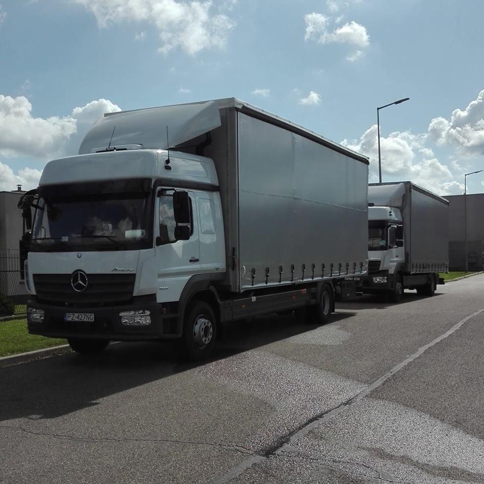 Dwa białe samochody ciężarowe Mercedes z szarą plandeką zaparkowane jeden za drugim na asfalcie w słoneczny dzień. Widoczna tablica rejestracyjna pierwszego pojazdu: PZ 427NG.