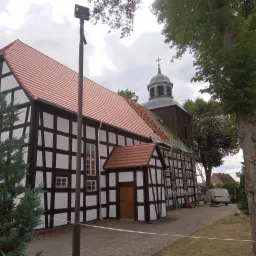 Kościół szkieletowy z czerwoną dachówką podczas prac remontowych, widoczne rusztowania i częściowo zdjęte stare pokrycie.