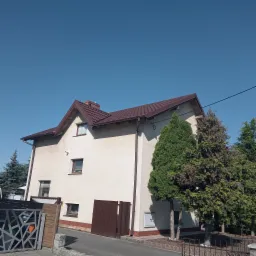 Dwukondygnacyjny dom z brązowym dachem z blachodachówki, widoczny z poziomu chodnika, z drzewami iglastymi w ogrodzie. Dach z rynnami, komin z cegły, okna z roletami.