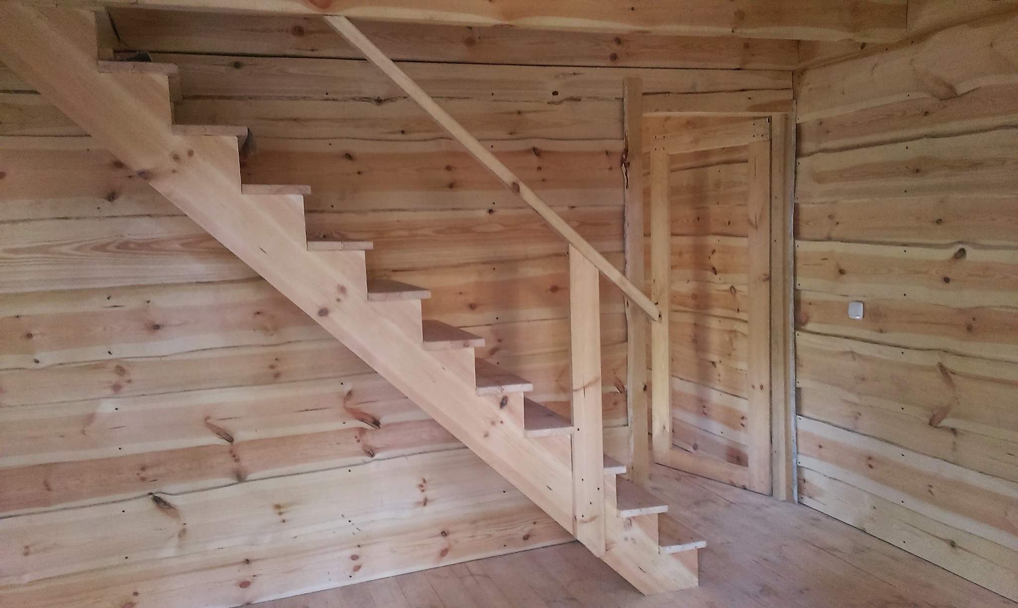 Surowe drewniane schody z prostą balustradą wewnątrz domu szkieletowego, widoczna drewniana ściana i drzwi.