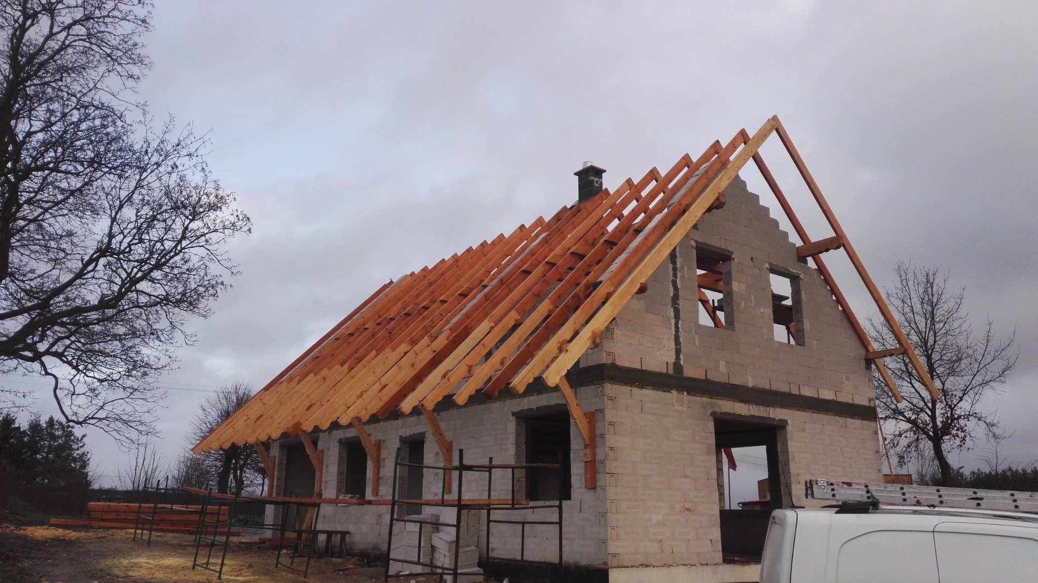 Surowy stan domu jednorodzinnego z białego bloczka, z częściowo wykonaną drewnianą konstrukcją dachu, widoczne rusztowanie i drabina oparta o samochód dostawczy na tle pochmurnego nieba.