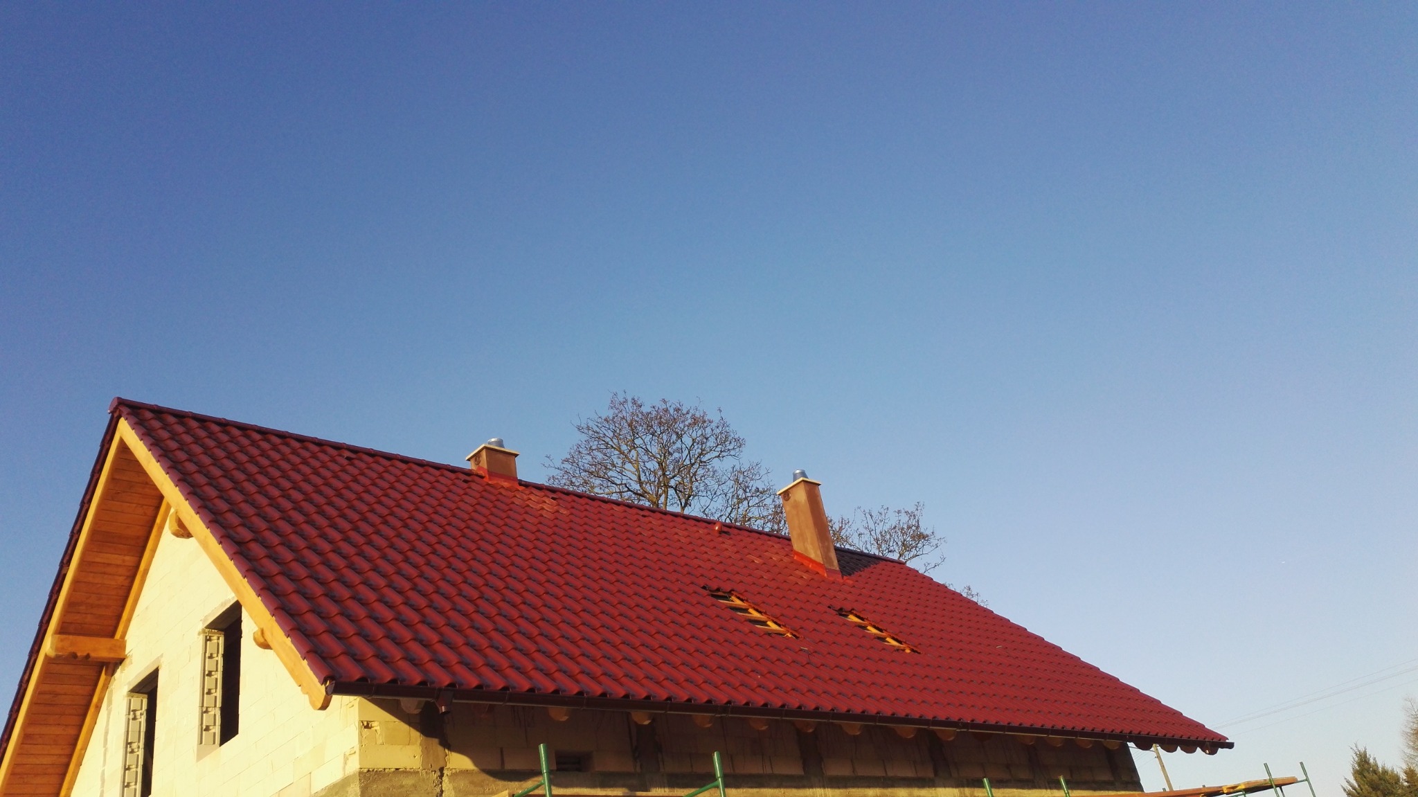 Czerwony dach z blachodachówki, dwa kominy i lukarny na tle błękitnego nieba i drzew bez liści, dom w trakcie budowy z widoczną więźbą dachową.