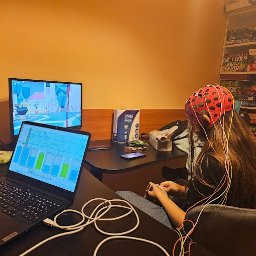 Dawid Milewski - Kobieta siedzi na krześle z elektrodami EEG na głowie, podłączona do komputera, na ekranie którego widoczne są wykresy aktywności mózgu, w tle półka z grami planszowymi.