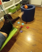 Kobieta rzuca woreczkiem do celu siedząc na huśtawce w pomieszczeniu z drewnianą podłogą, widoczne regały z zabawkami i niebieski puf.