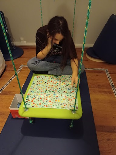 Dziewczynka siedząca na huśtawce z zielonym siedziskiem, układająca puzzle z drobnymi, kolorowymi elementami na podłodze z dywanem w tle.