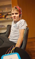 Chłopiec z elektrodami EEG na głowie i ręce, uśmiechnięty, siedzący w fotelu w gabinecie z półkami z książkami i grami planszowymi w tle.