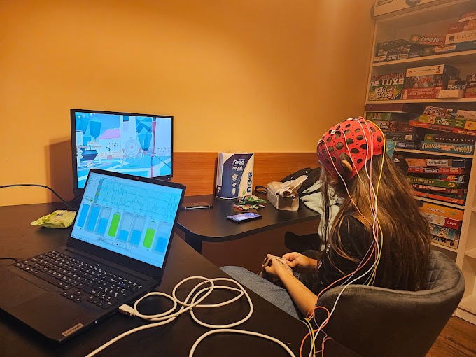 Kobieta siedzi na krześle z elektrodami EEG na głowie, podłączona do komputera, na ekranie którego widoczne są wykresy aktywności mózgu, w tle półka z grami planszowymi.