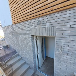 Zakład ogólnobudowlany Radosław Rozmarynowski - Szara ceglana elewacja budynku z drewnianą okładziną, betonowe schody prowadzące do otwartych drzwi wejściowych do piwnicy, widoczny fragment instalacji elektrycznej.