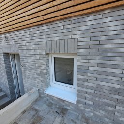 Zakład ogólnobudowlany Radosław Rozmarynowski - Elewacja budynku z szarej cegły klinkierowej z białymi oknami i drewnianym wykończeniem pod dachem, widoczne schody i narzędzie na posadzce.