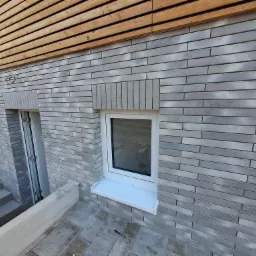 Elewacja budynku z szarej cegły klinkierowej z białymi oknami i drewnianym wykończeniem pod dachem, widoczne schody i narzędzie na posadzce.