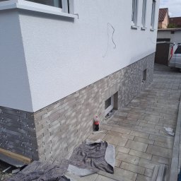 Zakład ogólnobudowlany Radosław Rozmarynowski - Elewacja budynku z szarej cegły klinkierowej, widoczne plastikowe krzyżyki dystansowe, przykryta plandeką, obok butelka Coca-Coli, kostka brukowa na podjeździe.