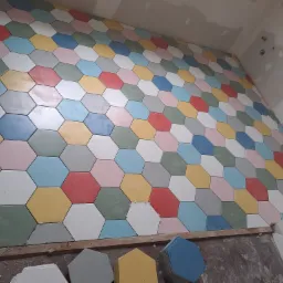 Ściana w trakcie układania z kolorowych, sześciokątnych płytek; widoczne płytki w odcieniach czerwieni, żółci, zieleni, szarości, błękitu i różu; na podłodze stosy płytek gotowych do montażu.