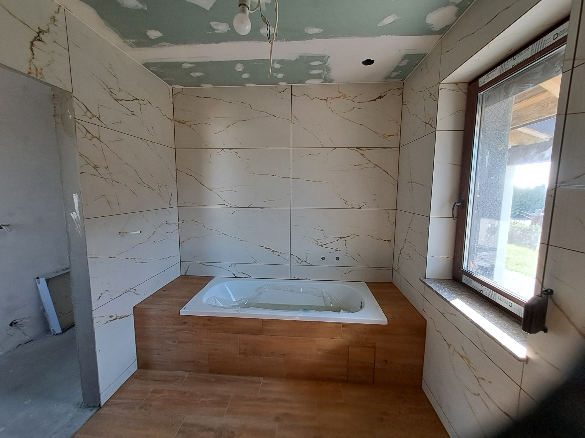 Niedokończona łazienka z wanną w obudowie z płytek imitujących drewno, ściany wyłożone płytkami z wzorem marmuru, surowy sufit z widocznymi śladami szpachlowania i wiszącą żarówką.