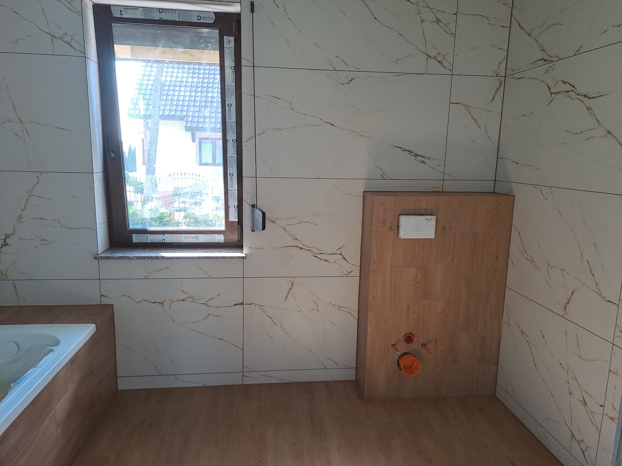 Ściana łazienki wyłożona białymi płytkami z brązowym wzorem żyłek, widoczne podłączenia do toalety podtynkowej, okno z widokiem na dom z szarą dachówką i fragment wanny obudowanej drewnopodobną...