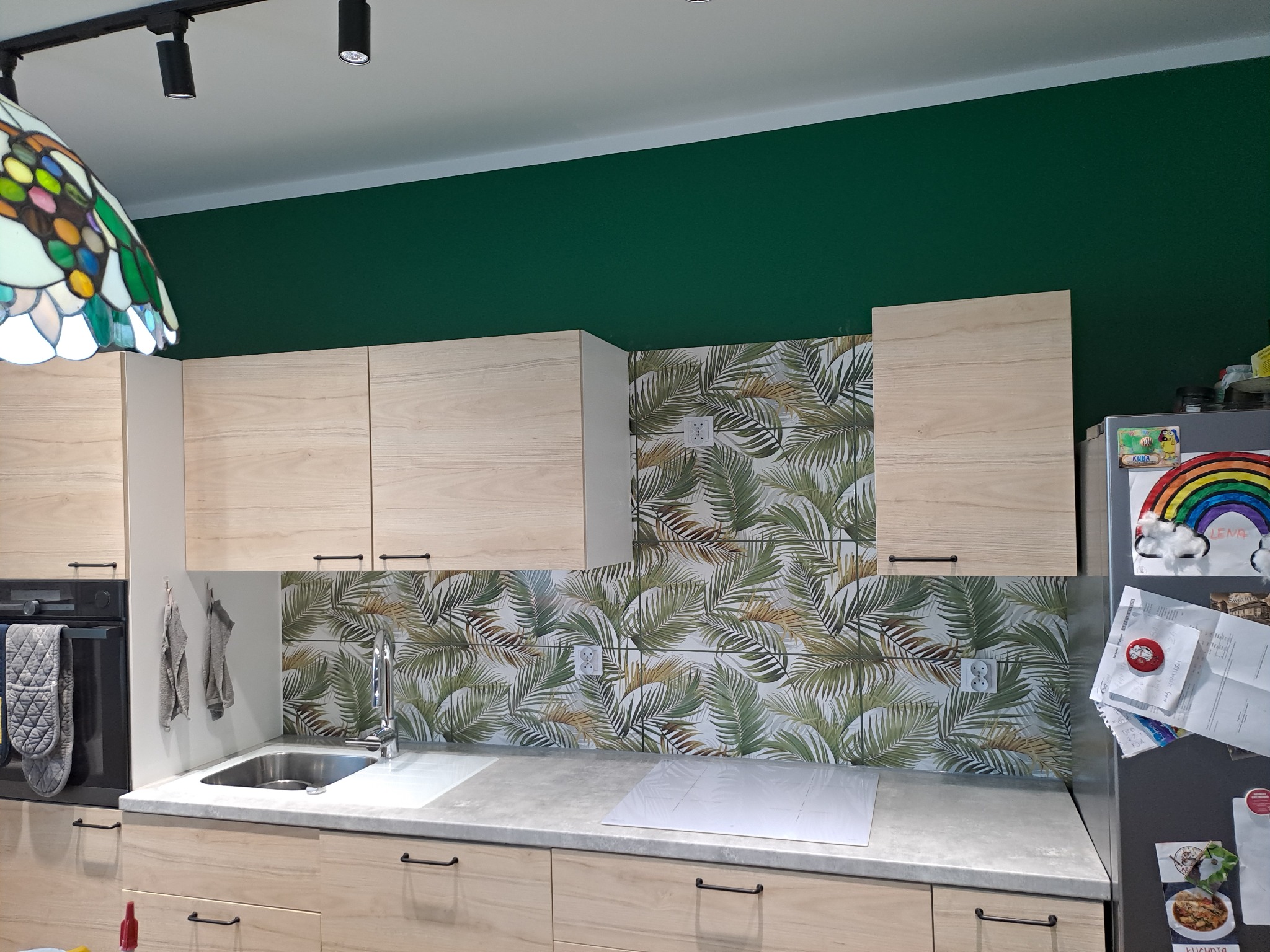 Jasna kuchnia z drewnianymi szafkami, blatem w kolorze betonu i płytkami z motywem liści palmowych na ścianie, widoczne gniazdka elektryczne. Nad zlewem wisi szary ręcznik, a na lodówce znajdują...