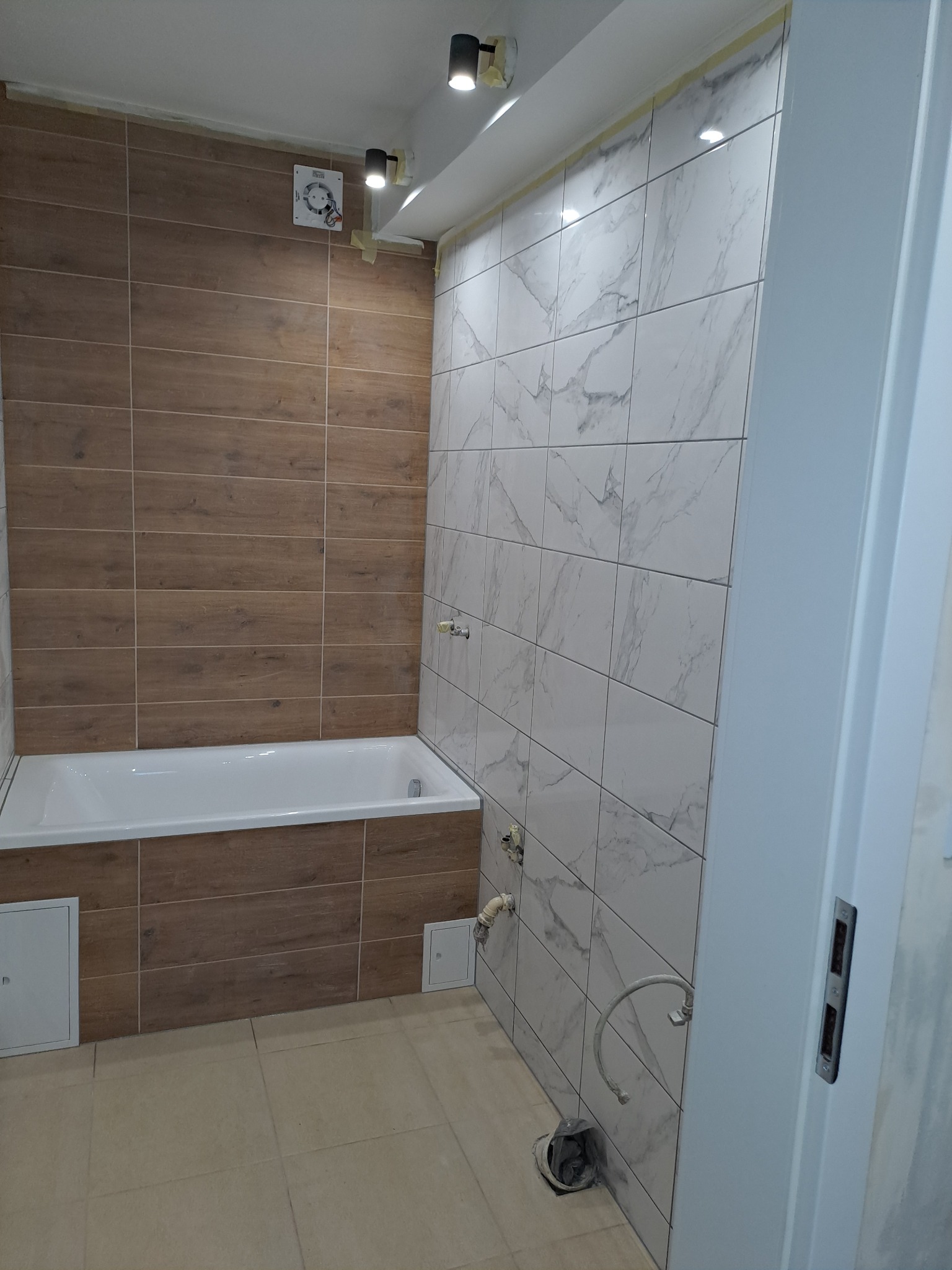 Wykończona łazienka z wanną obłożoną płytkami imitującymi drewno, ściana z białych płytek marmurkowych, widoczne przyłącza hydrauliczne i fragment drzwi.