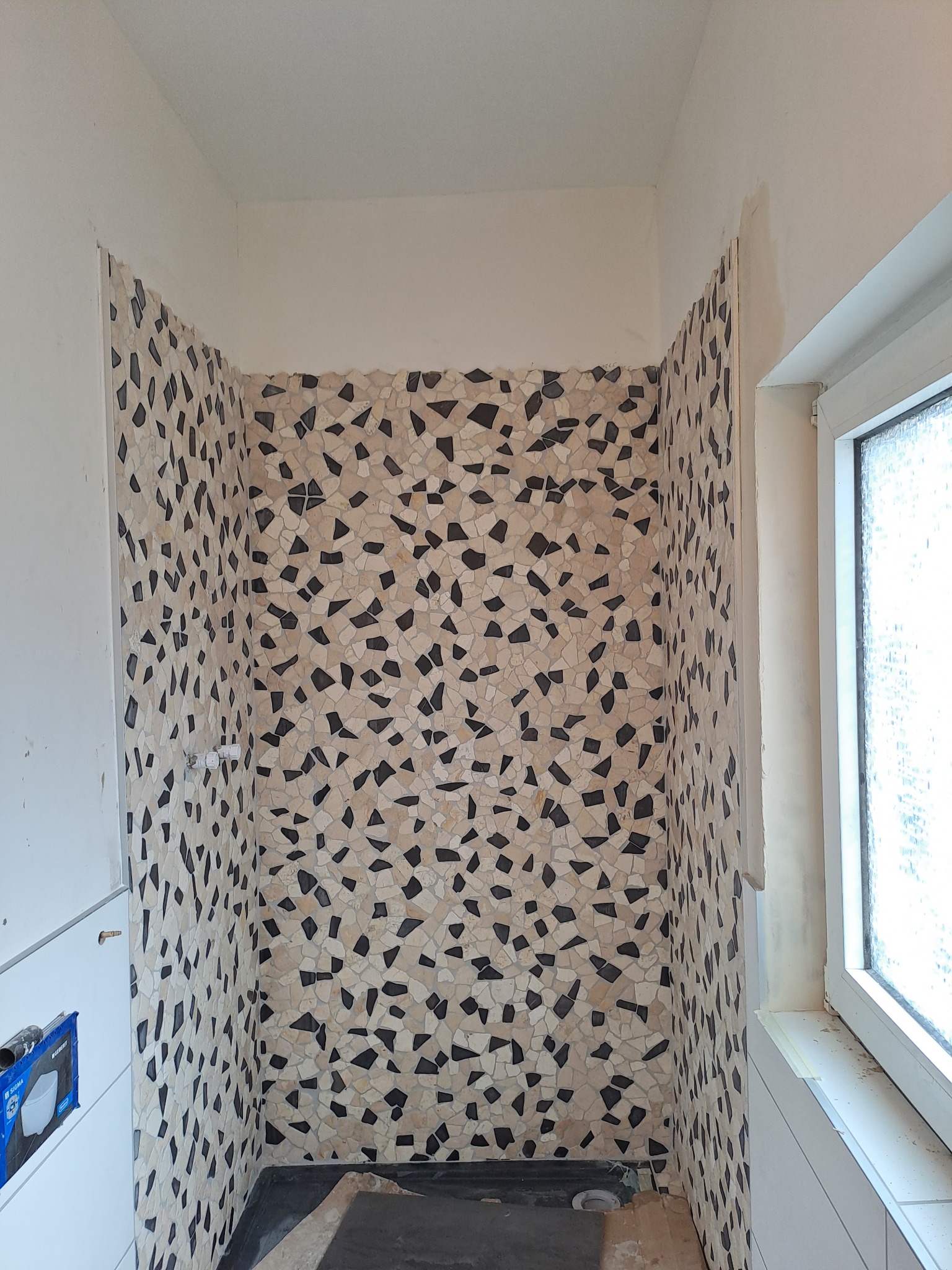 Fragment łazienki w trakcie prac wykończeniowych, ściany wyłożone mozaiką z nieregularnych elementów w odcieniach beżu i czerni, widoczny fragment okna i instalacji wodnej.