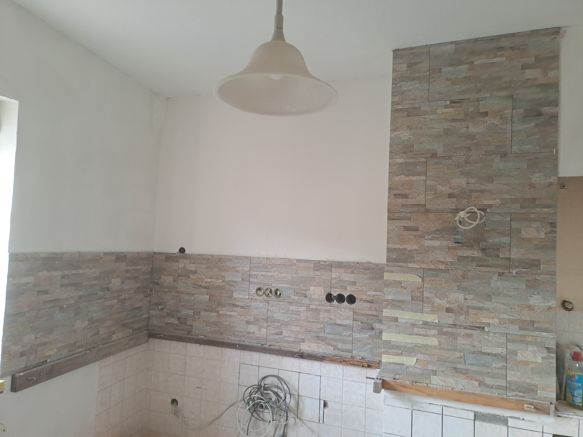 Kładzenie kamiennej okładziny na ścianie w kuchni, widoczne puszki elektryczne i przewody, dolna część ściany wyłożona białymi płytkami, fragment drewnianej listwy pod okładziną.