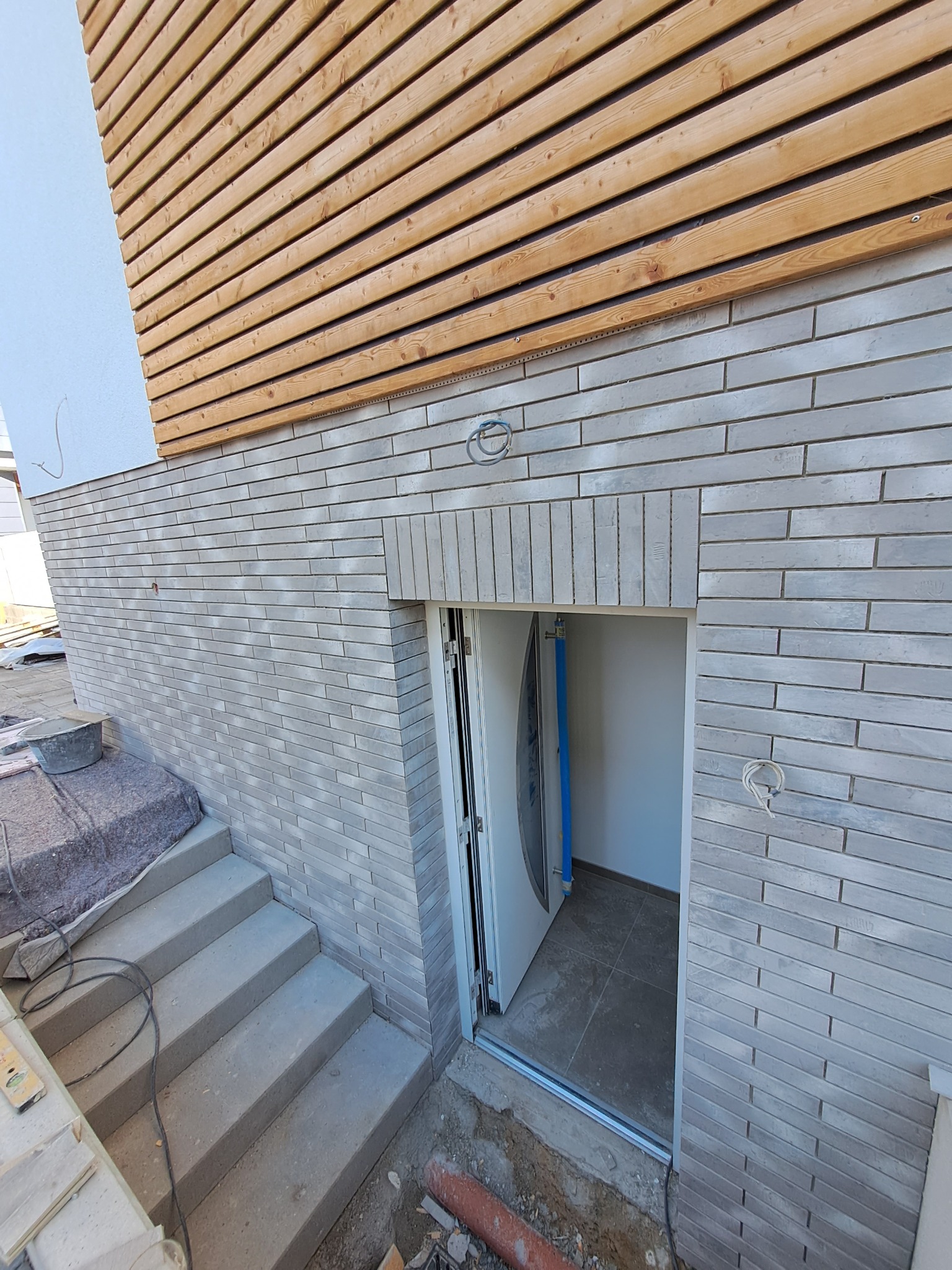 Szara ceglana elewacja budynku z drewnianą okładziną, betonowe schody prowadzące do otwartych drzwi wejściowych do piwnicy, widoczny fragment instalacji elektrycznej.