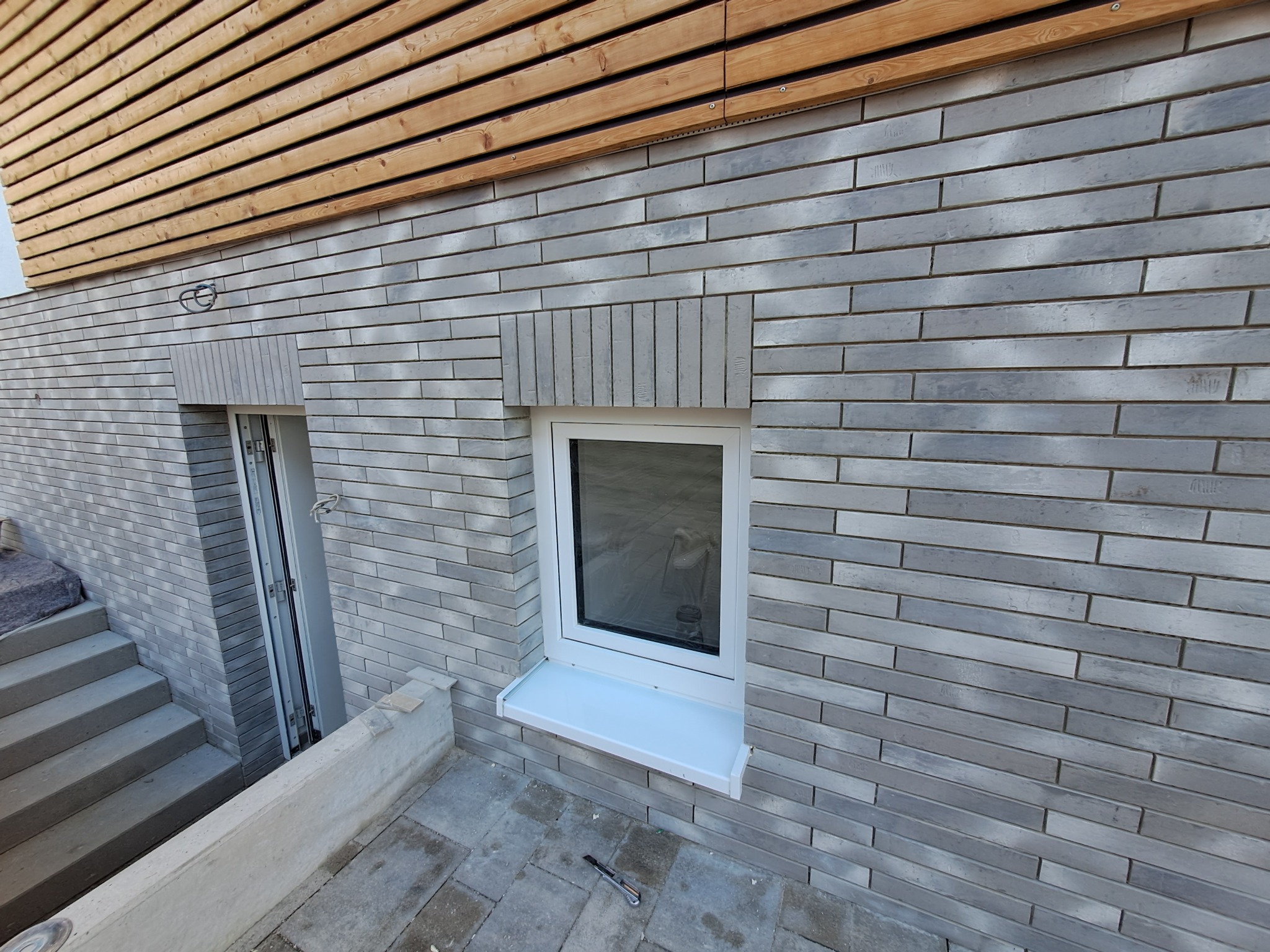 Elewacja budynku z szarej cegły klinkierowej z białymi oknami i drewnianym wykończeniem pod dachem, widoczne schody i narzędzie na posadzce.