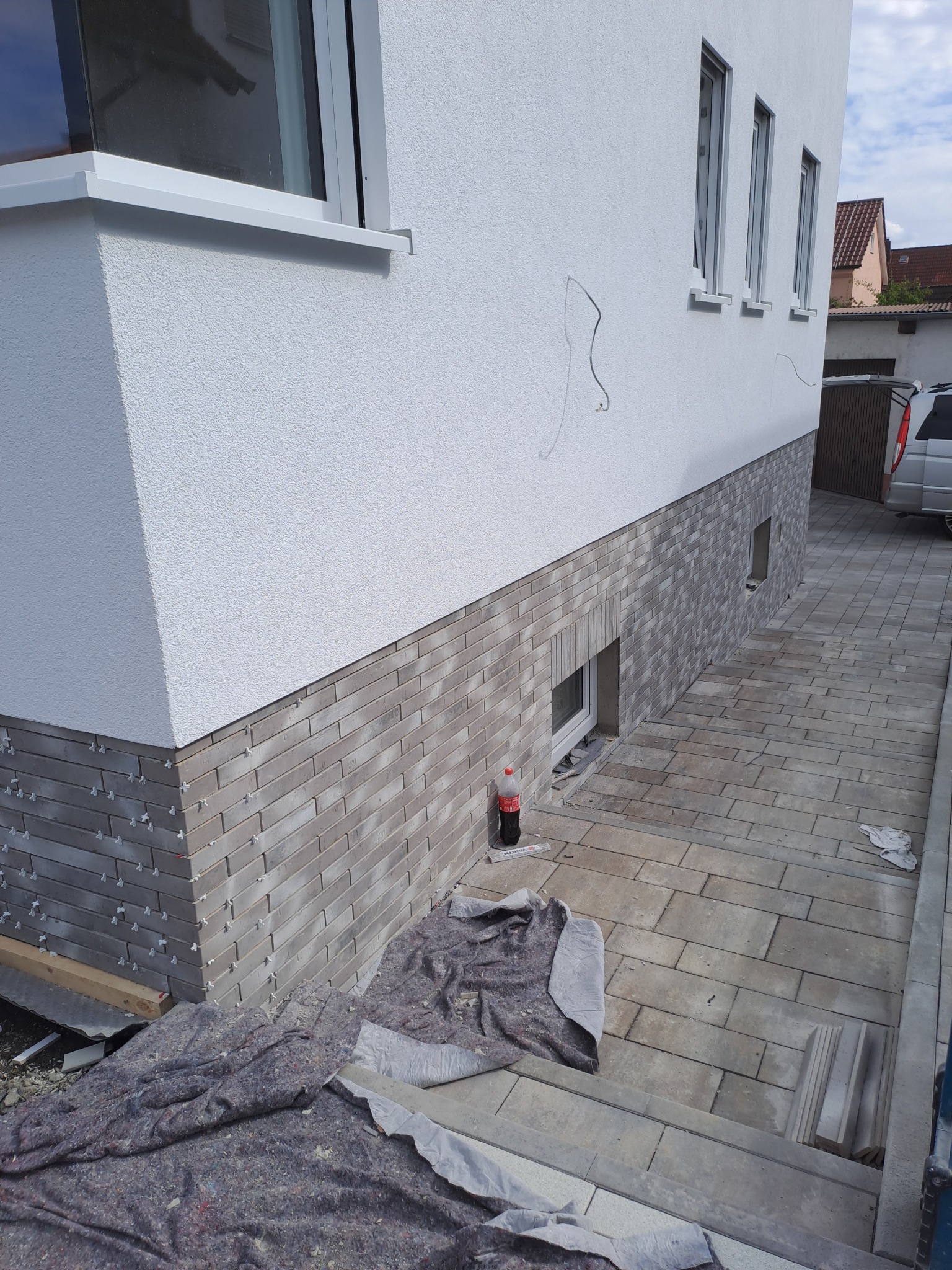 Elewacja budynku z szarej cegły klinkierowej, widoczne plastikowe krzyżyki dystansowe, przykryta plandeką, obok butelka Coca-Coli, kostka brukowa na podjeździe.