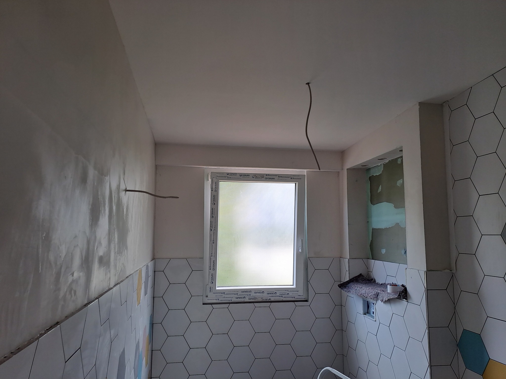 Wnętrze łazienki w trakcie prac: częściowo położone heksagonalne płytki, surowa ściana z widocznymi śladami szpachlowania, wystający kabel z sufitu, okno z widocznym logo producenta.