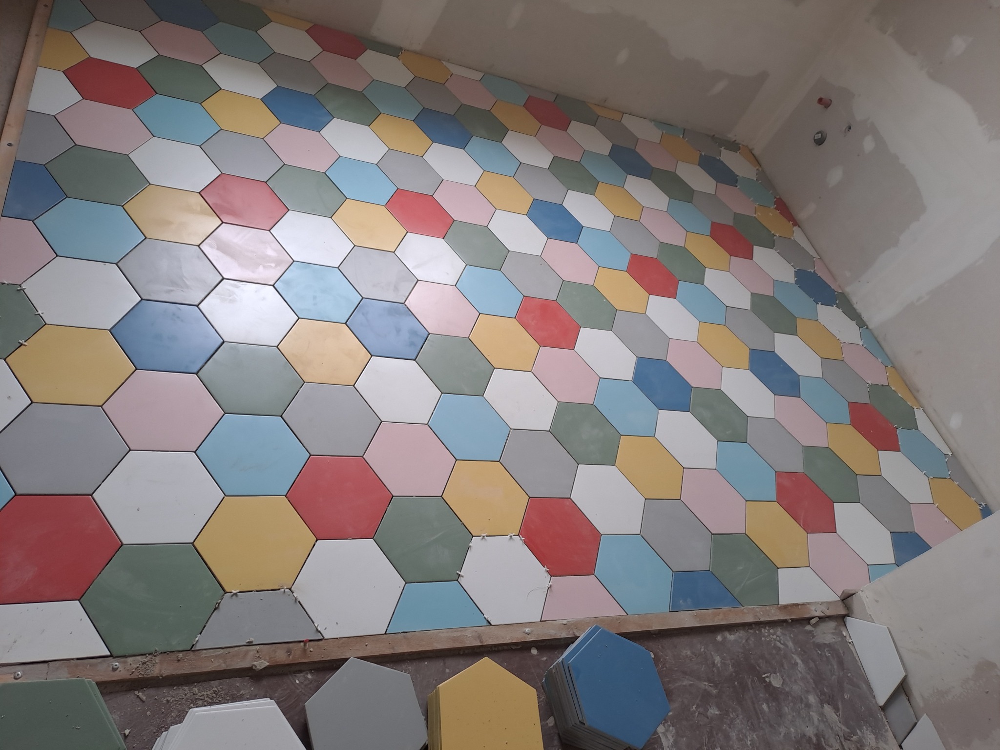 Ściana w trakcie układania z kolorowych, sześciokątnych płytek; widoczne płytki w odcieniach czerwieni, żółci, zieleni, szarości, błękitu i różu; na podłodze stosy płytek gotowych do montażu.