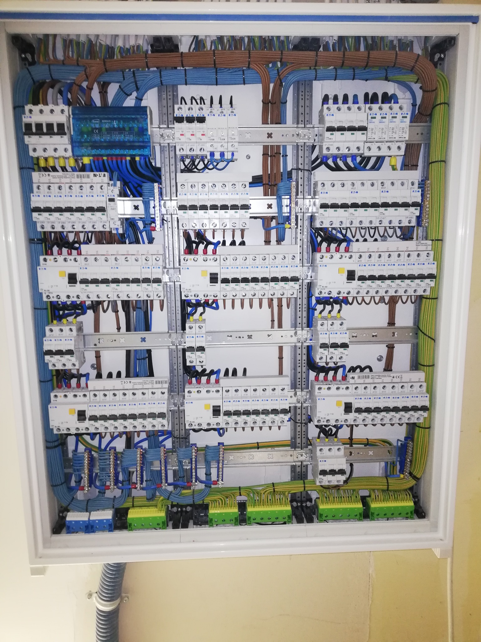 Rozdzielnica elektryczna z zainstalowanymi bezpiecznikami, przewodami w różnych kolorach (niebieski, brązowy, zielono-żółty), listwami zaciskowymi i szynami montażowymi, widoczne elementy marki Eaton.