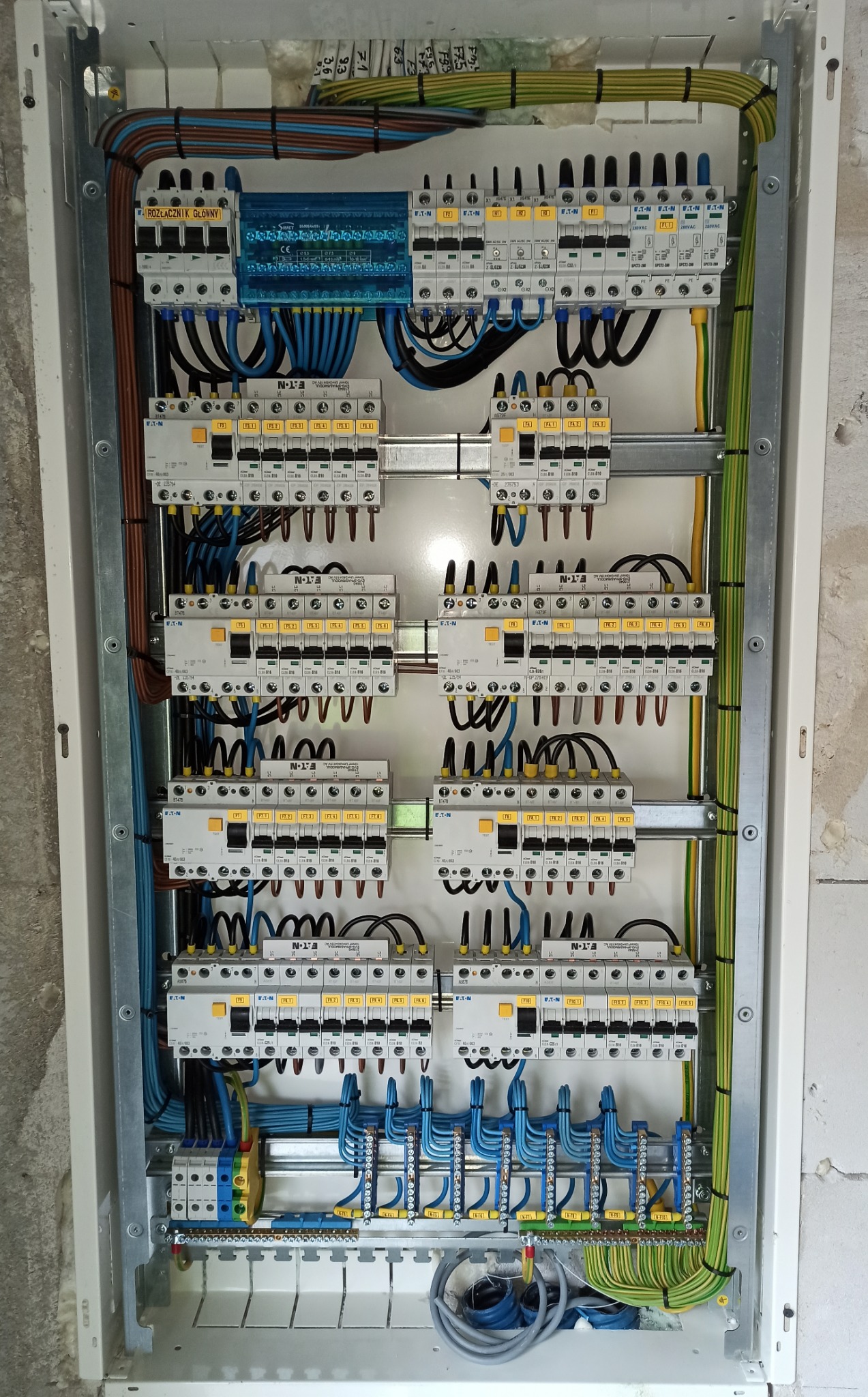 Rozdzielnica elektryczna w białej obudowie z widocznymi bezpiecznikami, przewodami różnego koloru i listwami zaciskowymi, zamontowana na ścianie w trakcie wykańczania.