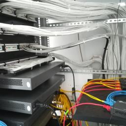 Msc Maciej Sampara - Szafa serwerowa z ułożonymi kablami sieciowymi, widoczne switche i patch panele, przewody w kolorach białym, żółtym, niebieskim i czerwonym.