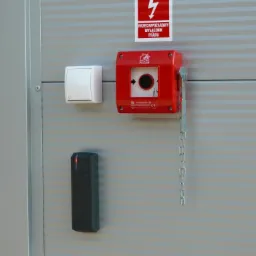 Przeciwpożarowy wyłącznik prądu w czerwonej obudowie z łańcuszkiem, biały włącznik światła i czarny czytnik kart dostępu zamontowane na szarej ścianie z poziomymi paskami.
