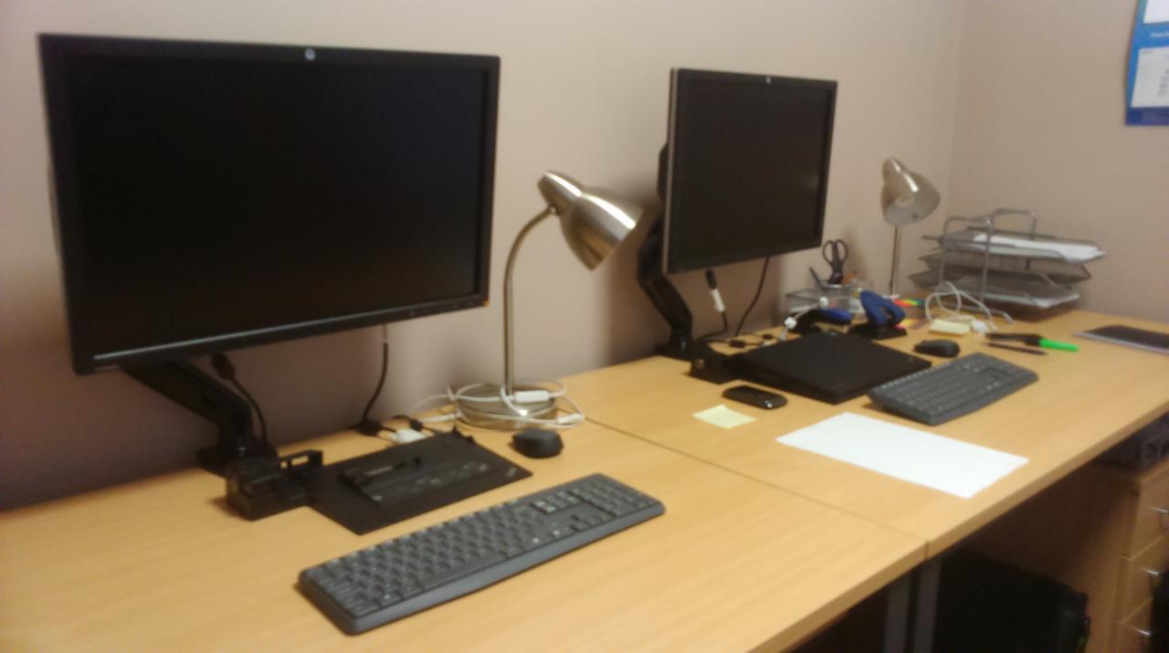 Dwa monitory na biurku z lampkami, klawiaturami, laptopem i organizerem na dokumenty, typowe stanowisko pracy.