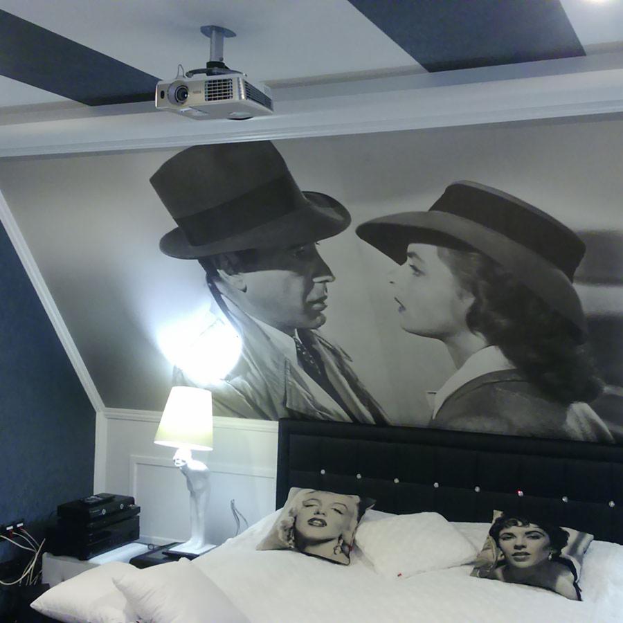 Sypialnia z projektorem sufitowym wyświetlającym czarno-biały mural filmowy, poduszki z wizerunkami ikon kina na łóżku, lampka nocna w kształcie figury.