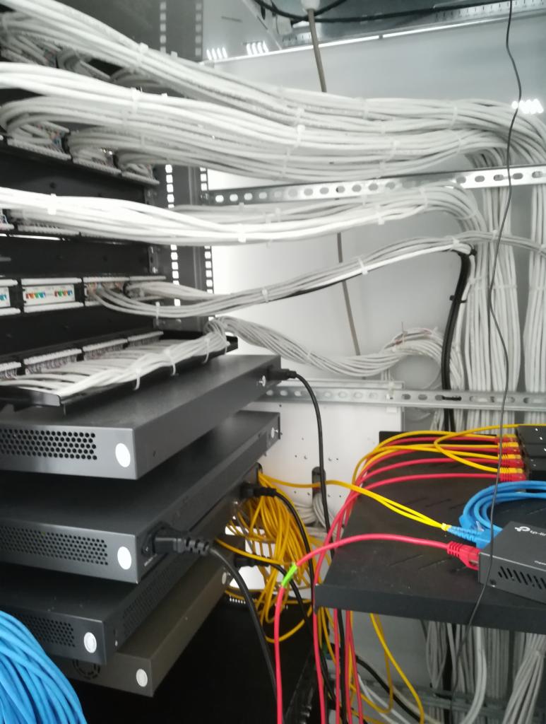 Szafa serwerowa z ułożonymi kablami sieciowymi, widoczne switche i patch panele, przewody w kolorach białym, żółtym, niebieskim i czerwonym.