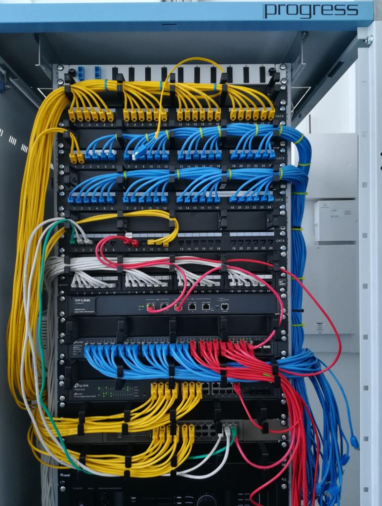 Rządowe okablowanie w szafie serwerowej z uporządkowanymi kablami Ethernet w kolorach żółtym, niebieskim i czerwonym, z widocznymi patch panelami i przełącznikami sieciowymi marek TP-Link i Pip-Link.