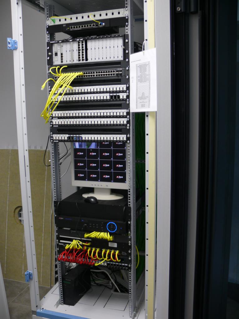 Szafa serwerowa z patch panelem, switchem, monitorem wyświetlającym ekrany kamer i okablowaniem w kolorach żółtym i czerwonym, widoczna dokumentacja techniczna po prawej stronie.